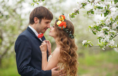 Wedding couplethe groom is a young darkhaired man in a black suit and pink bow tiebeautiful bridebrunette with long curly hair in a white wedding dress on her head a wreathposing embracing in the Park among the blooming trees.の写真素材