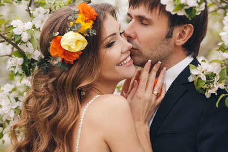 Wedding couplethe groom is a young darkhaired man in a black suit and pink bow tiebeautiful bridebrunette with long curly hair in a white wedding dress on her head a wreathposing embracing in the Park among the blooming trees.の写真素材