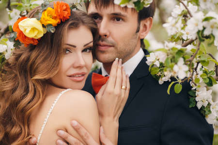 Wedding couplethe groom is a young darkhaired man in a black suit and pink bow tiebeautiful bridebrunette with long curly hair in a white wedding dress on her head a wreathposing embracing in the Park among the blooming trees.の写真素材