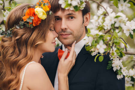 Wedding couplethe groom is a young darkhaired man in a black suit and pink bow tiebeautiful bridebrunette with long curly hair in a white wedding dress on her head a wreathposing embracing in the Park among the blooming trees.の写真素材