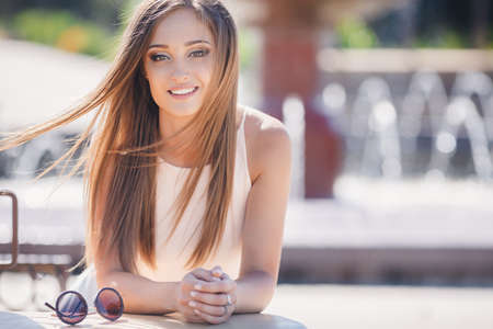Youngbeautifulattractive womana brunette with long straight hair and gray eyesa beautiful smile and straight white teethdressed in a white shirtsitting at a table in a city cafeposing against a green urban landscape.の写真素材