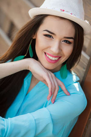 Brunette with long straight hair and brown eyesdressed in a shirt turquoise and blue jeansbeautiful smileearrings in your ears in the form of green leaves on his head a white hatposing sitting on the sidewalk near the wooden fence.の写真素材