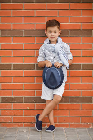 Stylish boybrunette with short hair on his head wearing a fashionable hat in dark greywearing a plaid light shirt on the neck under the shirt collar wearing a light grey scarfbaby posing on the street against the red brick wall in the summerの写真素材