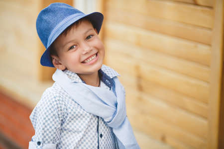 Stylish boybrunette with short hair on his head wearing a fashionable hat in dark greywearing a plaid light shirt on the neck under the shirt collar wearing a light grey scarfbaby poses on the background of a wooden house in the summerの写真素材