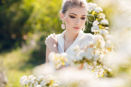 Young beautiful woman brunette hair is tied in her hairbig brown eyesstylish makeup in the ears earrings pinkdressed in a white sleeveless blouseposing in spring Parknext to a blossoming white flowers Bushの写真素材