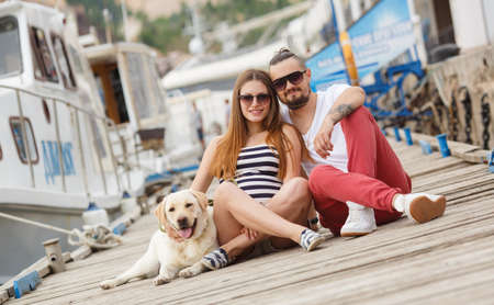 The happy couple, a man with a beard and a pregnant woman, a brunette with long straight hair, wearing sunglasses, with his friend Labrador dog, spend time on the pier near the yacht club enjoying the sea air and summer dayの写真素材