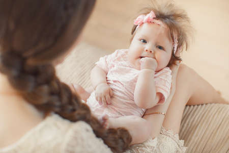 Portrait of a young happy family, mother is a beautiful woman, a brunette with long hair in a braid and grey eyes and her little daughter, a brunette with fluffy hair and gray eyes, posing in her room on a soft bed beigeの写真素材