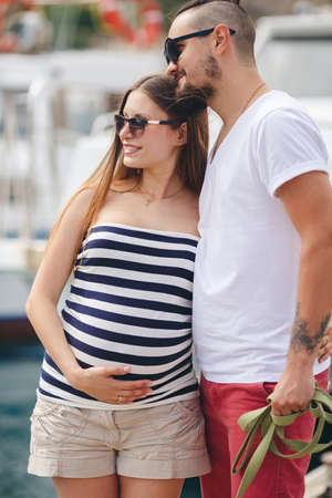 A young married couple, a man with a beard and a pregnant woman, brunette, long straight hair, spend their honeymoon together walking to the pier near the yacht club, enjoying the sea air and summer dayの写真素材