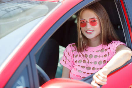 Happy young woman with a car .Young beautiful woman wearing glasses pink, brunette, long straight hair, dressed in a pink knitted t-shirt, rides around town in his red car in the summerの写真素材
