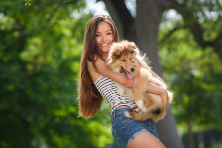 woman beautiful young happy with long dark hair in striped sweater holding collie dog. Young woman playing with Collie puppy outdoors in the park.の写真素材