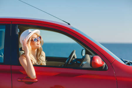 blonde beautiful woman in sunglasses and white hat sitting in red car by the sea. Sea view. Vacation...