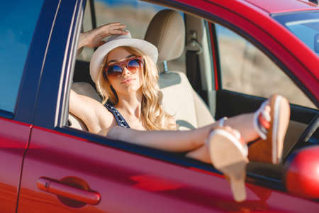 Beautiful blonde and red small car. Long-haired blonde in a white hat and striped dress, dark sun glasses, traveling alone on a small female red car on a rocky beach in the summerの写真素材