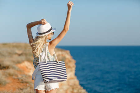 Blonde woman in summer hat and beach bag standing on the edge of the rock by the sea. Sea view. Freedom. Wind. Summer. Vacation. Yoga. looking to a sky and sea. Looking forwardの写真素材