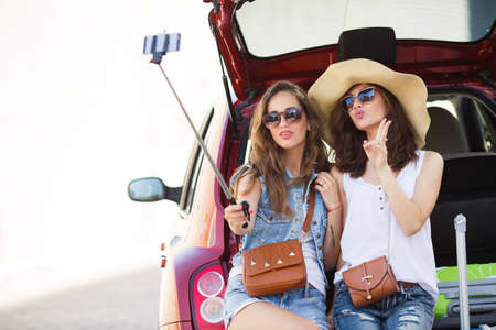 friends taking a selfie in the back of the car before leaving for vacations. Two women taking self portraits sitting in back car with suitcases. Vacation, trip concept. Road travel.の写真素材