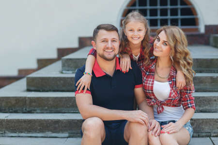 happy young family, father, mother and daughter spend the weekend summer day in the city park, parents and daughter look happy and smiling, happiness and harmony in family life, the family relaxing in the park sitting on the stepsの写真素材