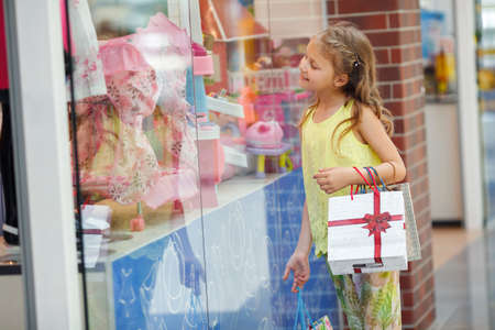 Smiling little girl with shopping bags in a large supermarket.Preschool age girl, blonde with curly hair in a city supermarket with paper bags, treats and shop windows, one without parents, wearing a light blouse and trousersの写真素材