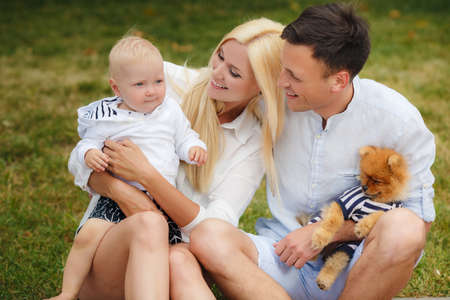 Happy young family, mother-blonde with a little red dog, dad, dark man with a young son, spend a day in a city park on the outdoors and having fun playing together on a background of green natureの写真素材