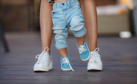 The first steps of the kid, Mom in white sneakers with a young son, dressed in blue pants and blue shoes, a warm summer day and learn to walk in the street, the first steps, the mother maintains her son.の写真素材