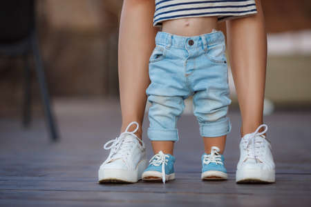 The first steps of the kid, Mom in white sneakers with a young son, dressed in blue pants and blue shoes, a warm summer day and learn to walk in the street, the first steps, the mother maintains her son.の写真素材
