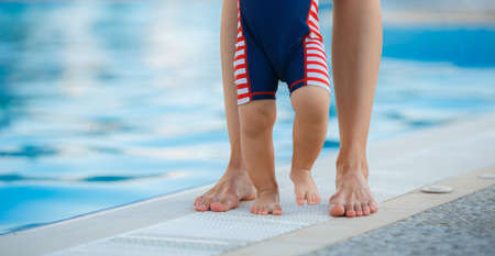 Legs of the child and mother after bathing in the pool, Beautiful legs of the mother and her baby, dressed in a striped bathing suit, standing by the pool with blue water in summer outdoors Mother and son walking beside the pool after swimmingの写真素材
