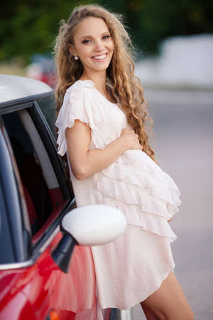 Cute young pregnant woman, a brunette with long curly hair and hazel eyes, dressed in a thin summer dress in white, beautiful smile, posing for the photographer standing near his favorite red car in the summer outdoors near the Parkの写真素材