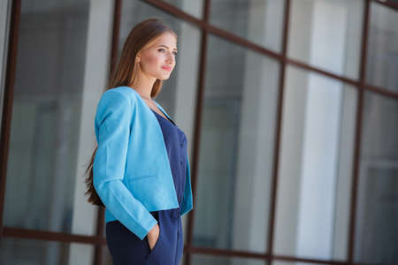 Morning portrait of a business lady, near the office.Portrait of a beautiful long-haired brunette with gray eyes dressed in a blue jacket, light makeup, brooding eyes, standing on the street near his bright office with large Windows during the breakの写真素材