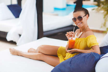 Young beautiful woman enjoying summer vacation on a tropical resort, a beautiful young woman in a yellow dress, holding a glass of cocktail while relaxing on a white lounger by the pool, beautiful, sexy woman drinking cocktailの写真素材
