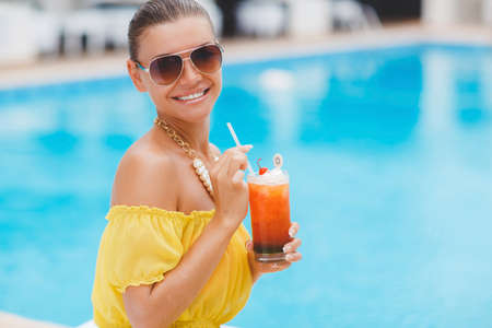 Young beautiful woman enjoying summer vacation on a tropical resort, a beautiful young woman in a yellow dress, holding a glass of cocktail while relaxing on a white lounger by the pool, beautiful, sexy woman drinking cocktailの写真素材