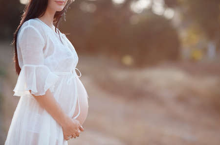 A pregnant woman waits boy, young pregnant woman, brunette, long straight hair with a big belly, dressed in a white transparent dress, hands on stomach, walking in the fresh air of the expectant mother expecting a sonの写真素材