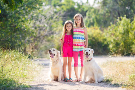 Two kids, two dogs, white Labradors outdoors in summer Park, girls, two sisters, playing with two big beautiful and good retrievers in the Park in the summer, children and dogs, happy familyの写真素材