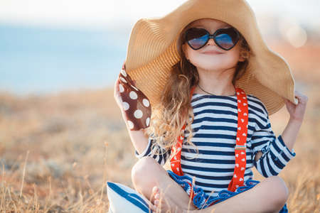 Happy little girl in a large hat, Beautiful young lady, a brunette with long curly hair, dressed in a striped sailor shirt and red suspenders, wearing dark sunglasses, sitting on a rocky beach in a big straw hat.の写真素材