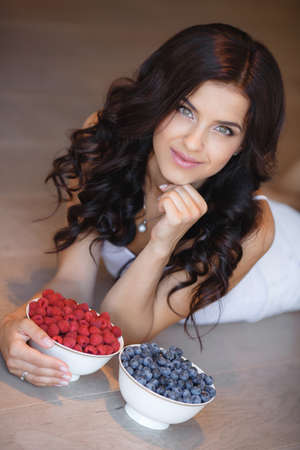 woman laying on the floor with bowl of berries blueberries at home. fresh morning. healthcare. Portrait of beautiful young woman eating a bowl of blueberries and smiling. Healthy food.の写真素材