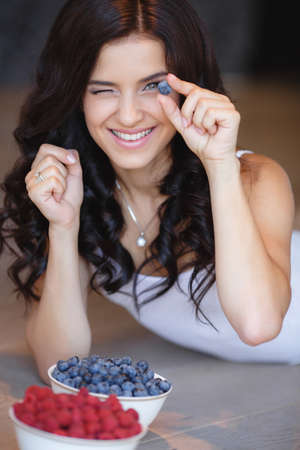 woman laying on the floor with bowl of berries blueberries at home. fresh morning. healthcare. Portrait of beautiful young woman eating a bowl of blueberries and smiling. Healthy food.の写真素材