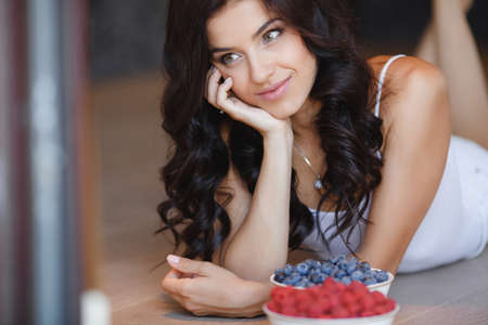 woman laying on the floor with bowl of berries blueberries at home. fresh morning. healthcare. Portrait of beautiful young woman eating a bowl of blueberries and smiling. Healthy food.の写真素材