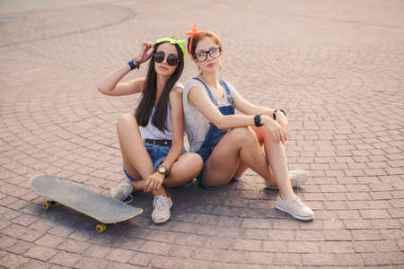 Two beautiful girlfriends, young athletic girl brunette in sports shoes and shorts, glasses, ride on a skateboard in the city, sunny, leisure, portrait of a smiling woman outdoors, healthy lifestyle, extreme sports.の写真素材