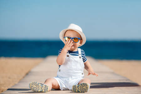 Portrait of the happy child in a hat on the seashore, Funny smiling child in a straw hat, a white summer suit and a striped shirt plays alone on a sandy beach against the blue sea and blue skyの写真素材