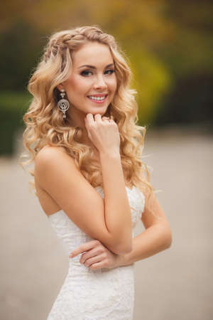 Bride in wedding dress posing for a photograph in the Park in the summer, Very beautiful woman in wedding white dress, with long, curly, blond hair, blue eyes, beautiful smile and beautiful earrings, posing in summer green park.の写真素材