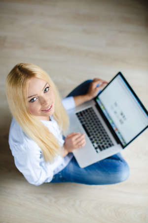 Happy young beautiful woman the blonde with blue eyes and long straight hair, light makeup, nice smile, white teeth, dressed in a fashionable white shirt and blue jeans, conducts correspondence in social networks using laptops, sitting in the house on theの写真素材