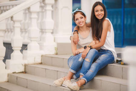 Lifestyle, two beautiful young women, beautiful girlfriends, brunette, dressed in a light sleeveless shirts and blue jeans, spend time together on city streets in the summer, having fun and smilingの写真素材