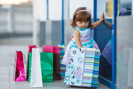 A small child, a girl, a brunette with curly hair and brown eyes, in a light summer dress, one is near a shopping Mall with colorful bags, cute baby goes shopping with bags, shopping little girl in the big city.の写真素材