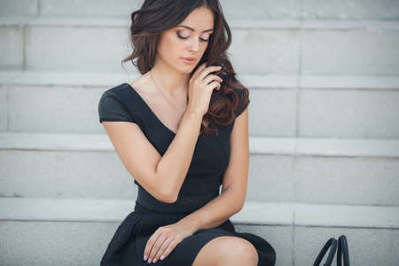 A beautiful young woman of Asian appearance, with long brunette curly hair, in a black short dress without sleeves, a student of the prestigious educational institution, sits on the steps near the white railing, summer, Sunny dayの写真素材