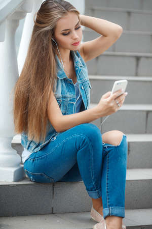 A lovely young woman, brunette with long straight hair and gray eyes, dressed in a dark blue denim jacket without sleeves, sitting on the stairs in a subway, wearing headphones, listening to your favorite music from your mobile phoneの写真素材