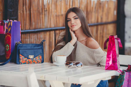 Young beautiful brunette woman with long straight hair and brown eyes, after an exhausting shopping with colored bags, spends his time sitting at a table in a summer cafe with a cup of hot coffee.の写真素材