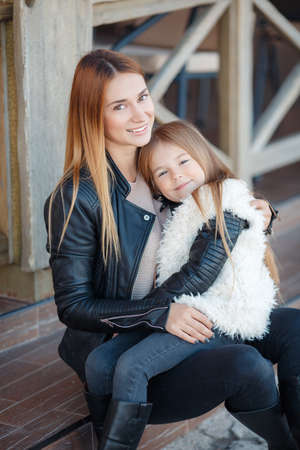Portrait of happy family, mother and daughter, daughter brunette and redhead mom, long straight hair, dressed in black leather jackets, spend time together, sitting together on the steps outside the house, Sunny spring day, joy and family happinessの写真素材