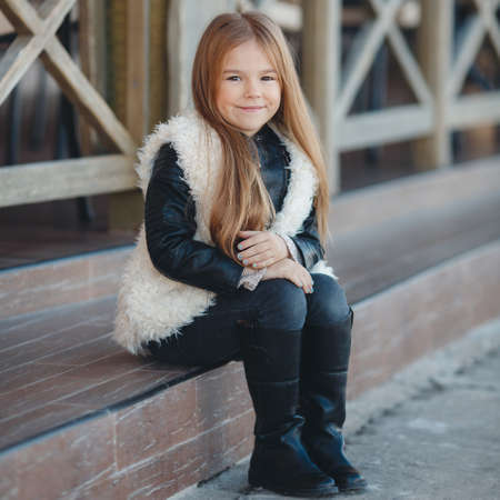 Little girl sitting on the steps near the house.Cute preschool girl, brunette with long straight hair and brown eyes, wearing a black jacket and a white fur vest, resting at his dacha outside the city, sitting on the steps near the houseの写真素材