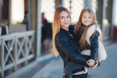 Spring portrait of a happy family, red-haired mother and daughter brunette with long straight hair, dressed in black leather jackets, spend time together, mother holds daughter on hands standing near a country house.の写真素材