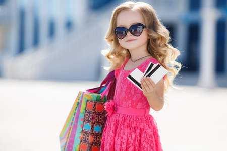A beautiful little girl of preschool age with a credit card, a brunette with curly hair, pink summer dress, colorful paper bags, sunglasses, with a sweet smile, one goes shopping to the Mall, shopping, mom's helper in the big cityの写真素材