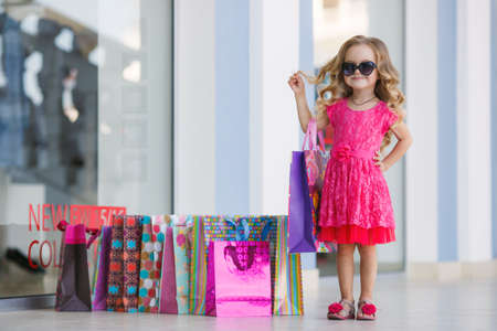 A beautiful little girl of preschool age, a brunette with curly hair, pink summer dress, colorful paper bags, sun glasses, with a sweet smile, one goes shopping to the Mall, mom's helper in the big city, shoppingの写真素材
