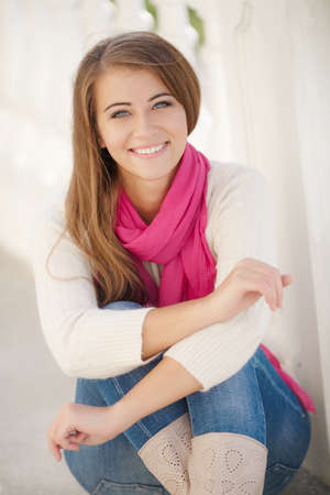 Happy young woman brunette with long straight hair and gray eyes, spends time outdoors in the city, dressed in a white sweater, jeans and pink scarf, a beautiful smile, posing for the photographer on the street in the afternoonの写真素材