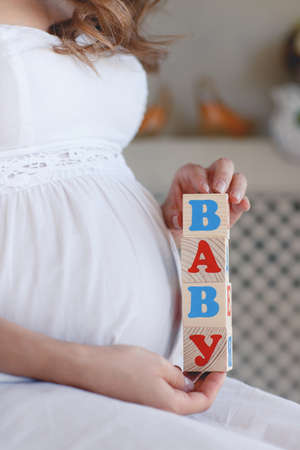 The pregnant woman with toy cubes in hands.Beautiful, young, pregnant woman, brunette with long curly hair, sitting in her room, holding the wooden pieces and puts them into words boy and babyの写真素材